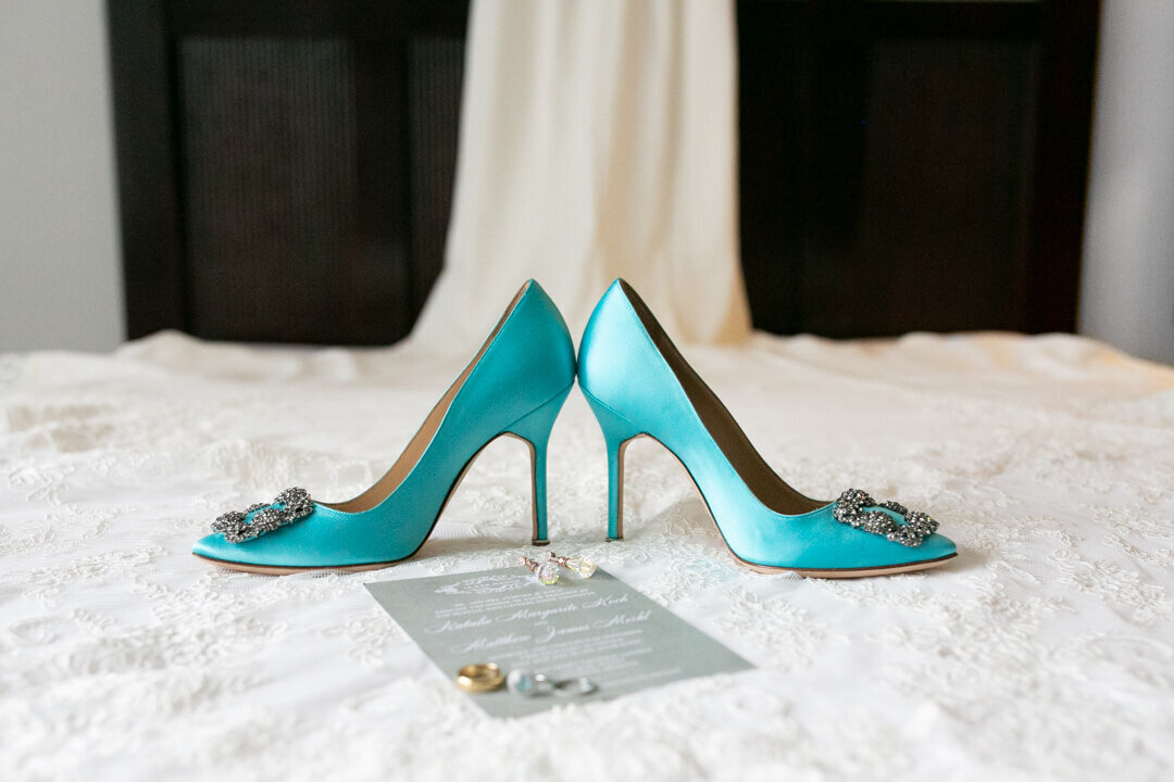 Turquoise bridal shoes placed with invitation and rings on top of dress taken by Photography by Karla.