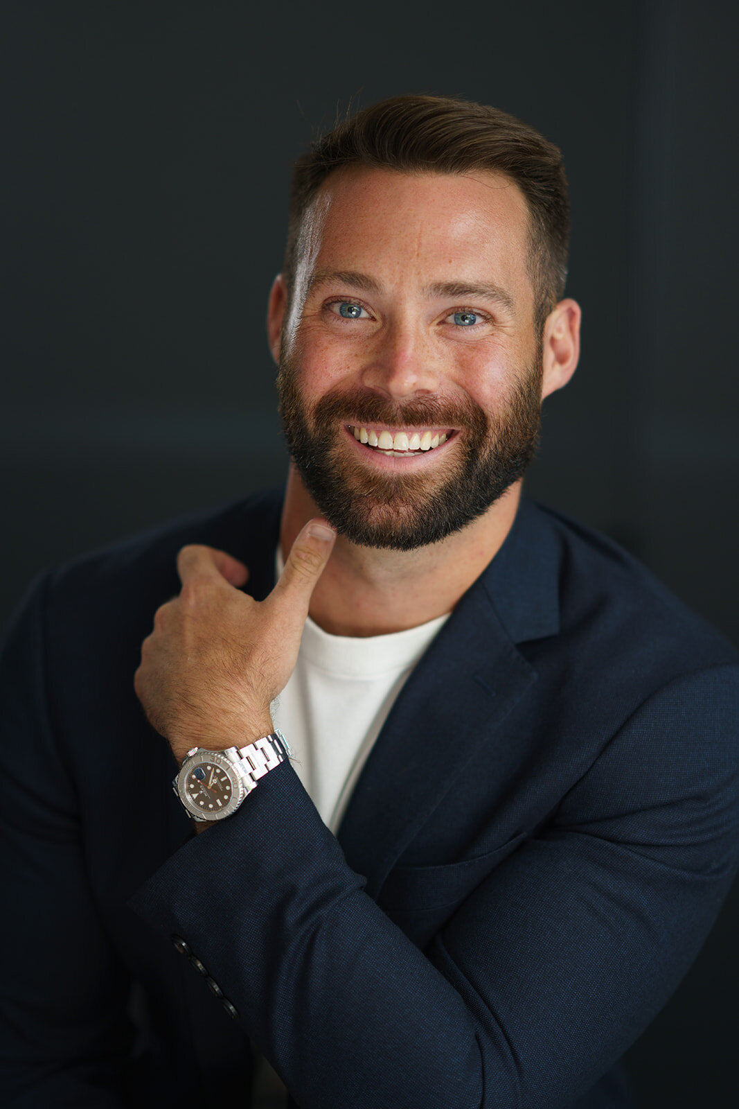 Dramatic close-up headshot of a young, bearded man smiling while showing off his watch and wearing a dark suit against a dark background.