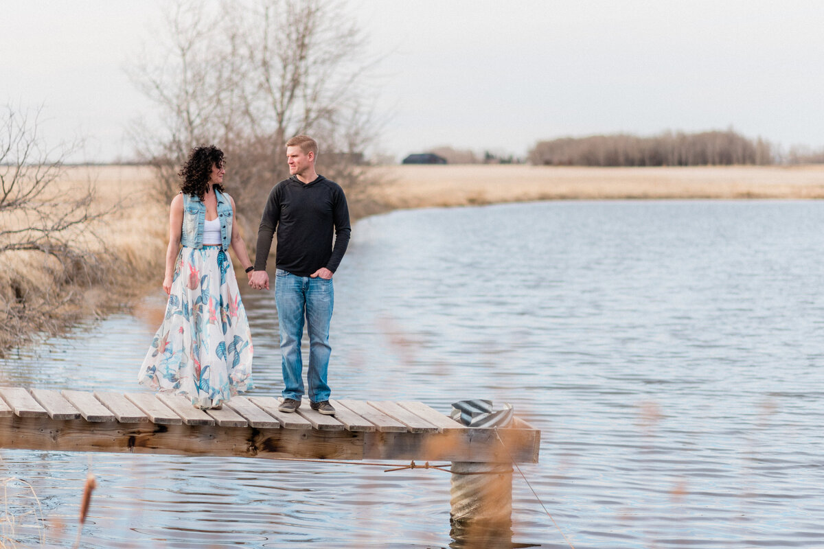 Couple holds hands and gazes at each other while standing on a dock over water.
