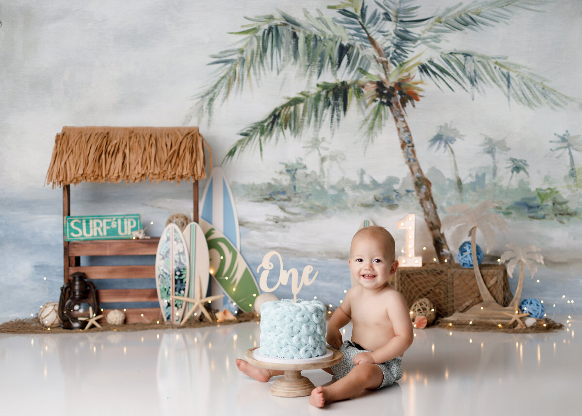 Surf and beach themed cake smash in West Palm Beach and Loxahatchee Florida studio. Baby is sitting behind a baby blue cake with a swim trunks on. In the background is a cute tiki hut with surfboards, palm trees and beach theme accents.