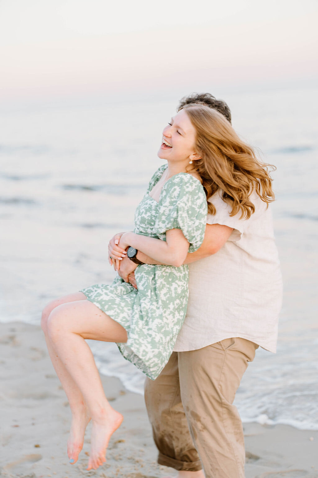 Newport Rhode Island Engagement Photo Photographer | A couple enjoys a playful moment by the beach. The woman, in a green floral dress, laughs as she's lifted. 