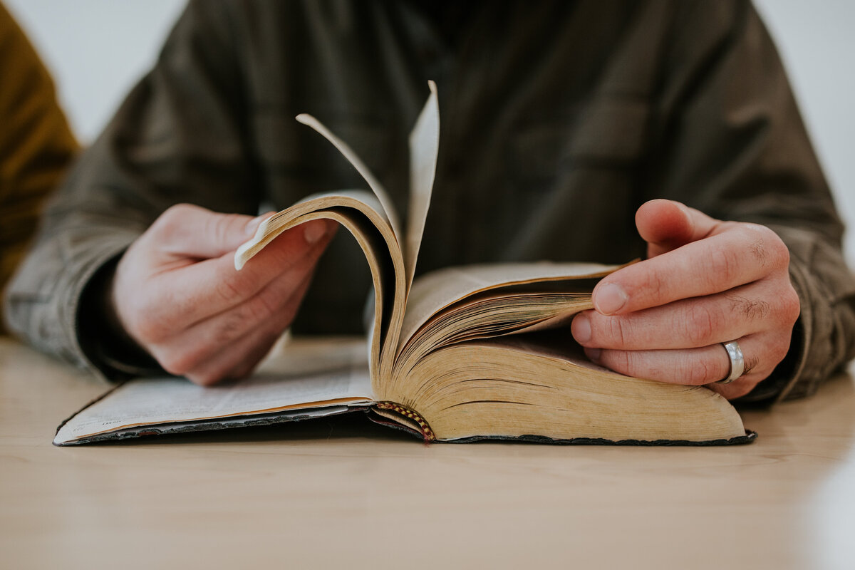 Camera looks at man flipping through Bible on table.