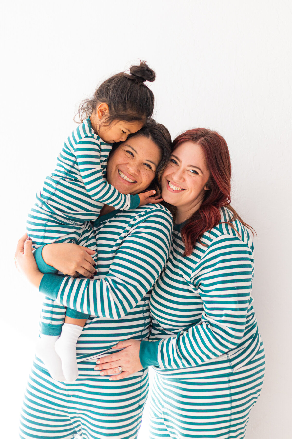 two moms in hug their daughter. they wearmatching pajamas against a white background at a los altos photography studio