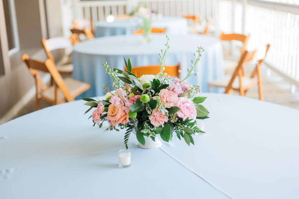 A round table with a light blue tablecloth features a floral centerpiece of pink and peach flowers and greenery, with a small white candle beside it. Wooden chairs and similar tables are in the softly lit background.