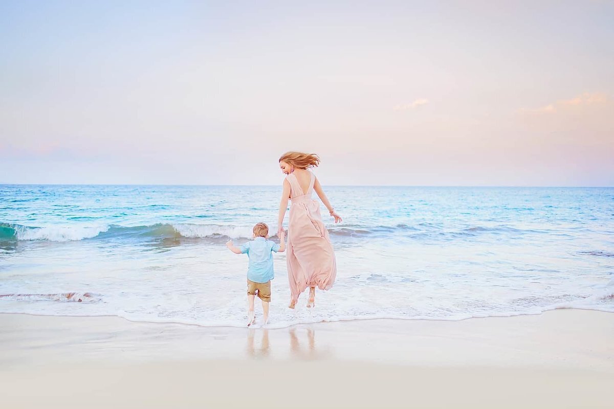 Candid family portrait of mom holding son's hand on the beach in Maui as she jumps and he runs toward the water