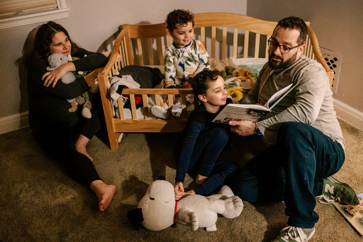 Family of four gathered around and inside a toddler bed in the dim light, as the father reads from a Dr. Seuss book. Kids are in their pajamas, and parents look tired but content.