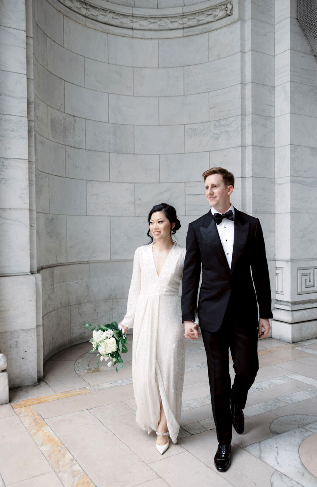 The bride and the groom are holding hands inside New York Public Library, NYC, with a very high ceiling. Image by Jenny Fu Studio