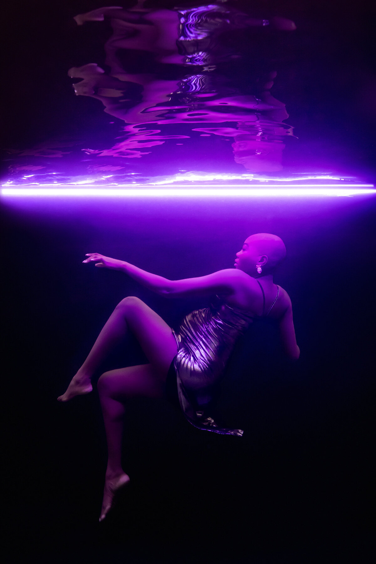A striking underwater portrait captured by Lindsay Walden Photography showcasing a woman gracefully suspended in water against a deep black backdrop. Illuminated by a vivid purple neon light, her metallic gown shimmers beautifully, highlighting the luxurious, ethereal feel of the scene. Dallas maternity photographer masterfully blending dramatic lighting and elegance.