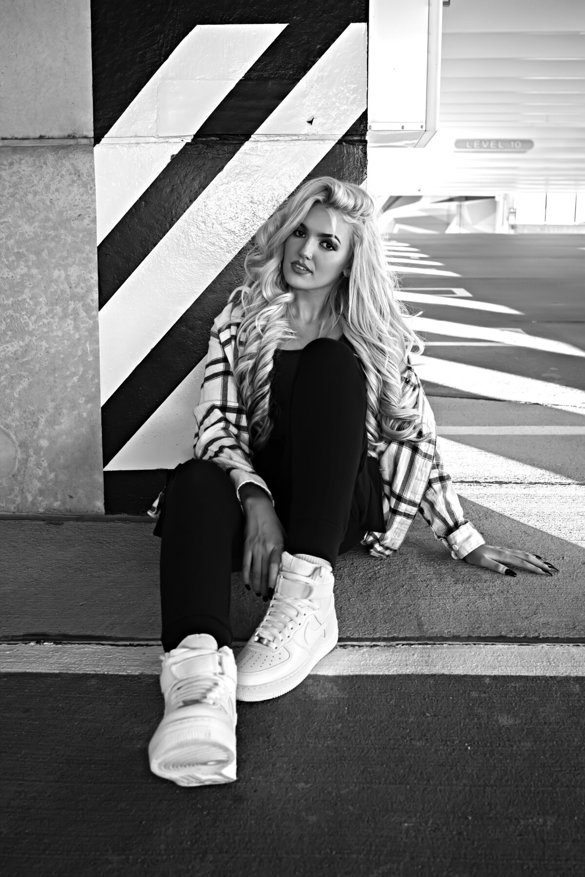 Woman in white, black, and lime green plaid shirt, black tank top, black joggers, and white Air Force 1s sitting on ground in a parking garage