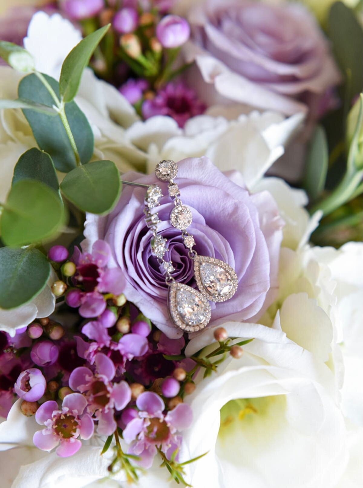 Dangling diamond earrings layingon top of a light purple rose  in a bridal bouquet of white hydrangea, eucalyptus and purple wax flower.