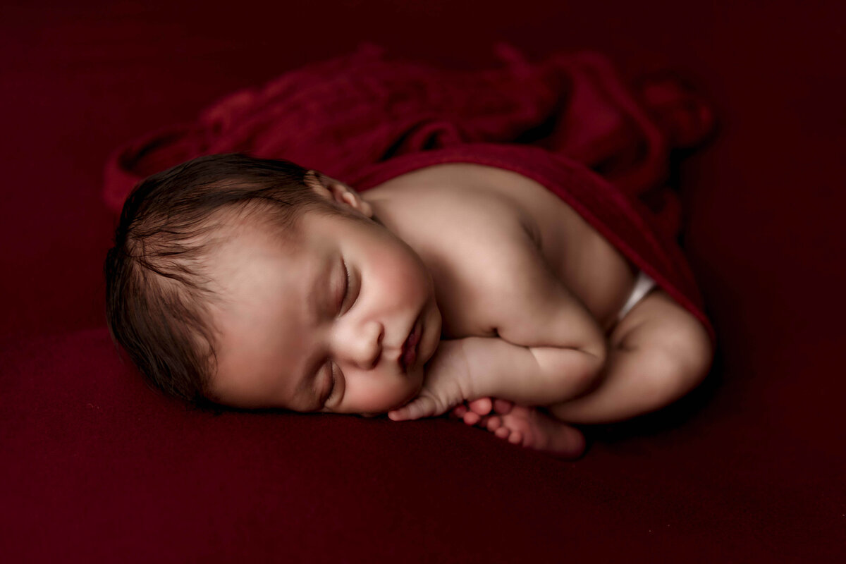 A gorgeous fine art newborn photo by Mobile, AL newborn photographer. This baby is sleeping peacefully in the taco postion, with his feet carefully folded underneath him much like how a baby is in the womb. He has a head full of dark hair, and is resting on a deep red beackdrop that perfectly compliements his skin tone and hair.