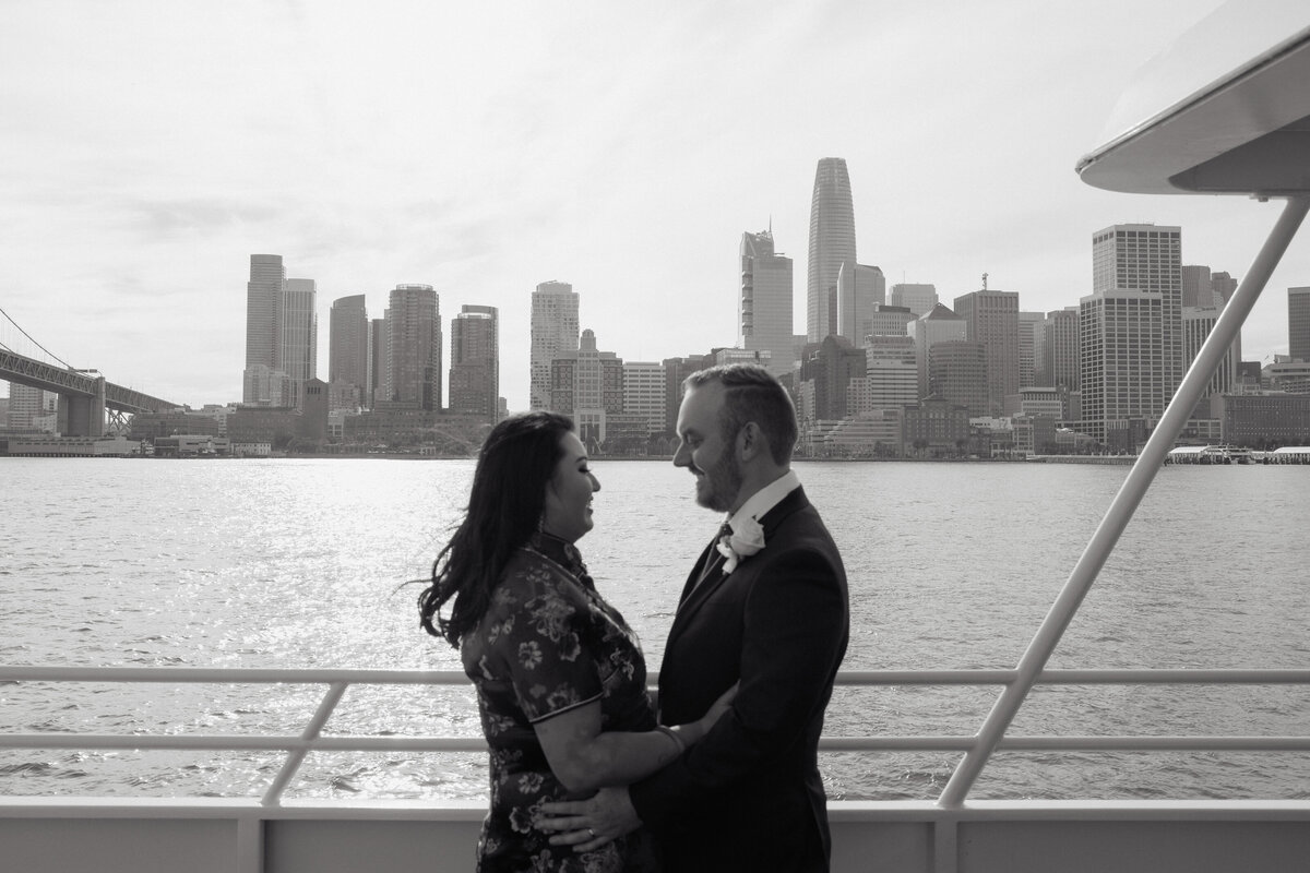 Bride and groom looking lovingly at each other aboard yacht with San Francisco skyline in background, candid wedding photo.