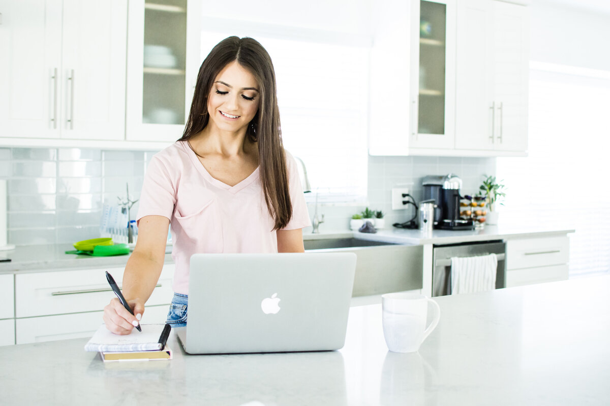 Woman with dark hair works at kitchen counter with laptop and notebook