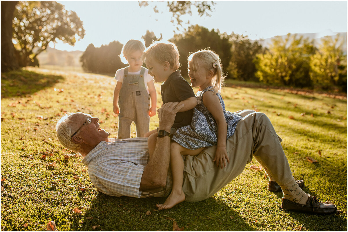 Granddad lying on his back on the grass, playfully letting two children ride on him like a train.