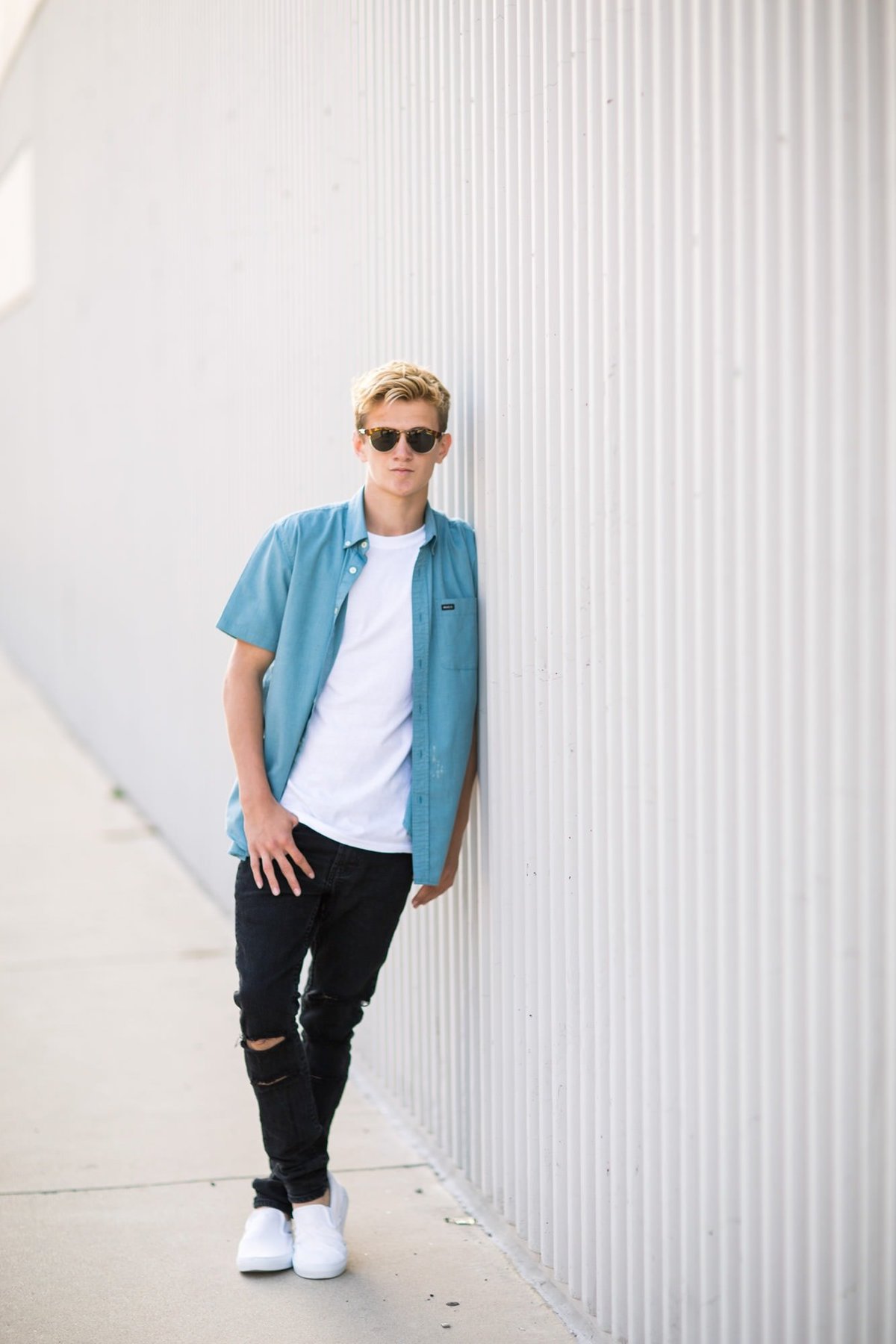 High school senior boy leans against a wall posing with his sunglasses on