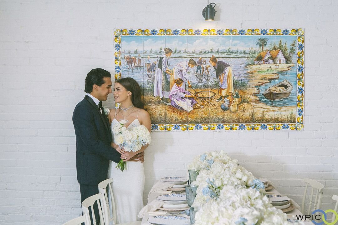 a bride and a groom standing next to each other beside a spanish inspired mural and behind a fancy wedding reception table filled with white flowers