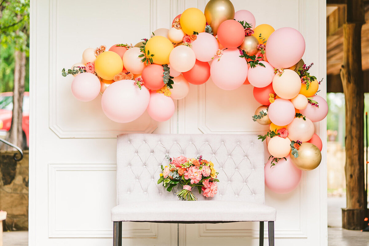 White couch sitting underneath a pink, orange and gold balloon arch decorated with greenery