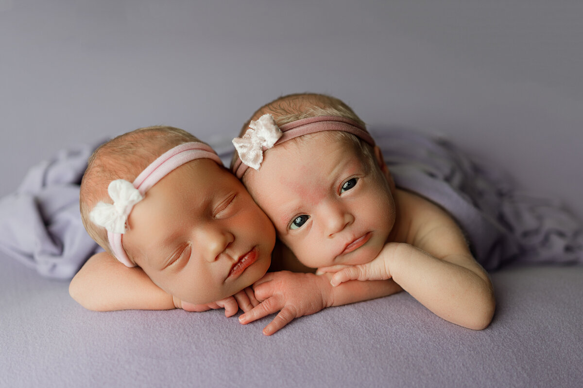 twin newborn girls posed together laying forward on tummy with hands under chin on a purple backdrop. a proffessional photo