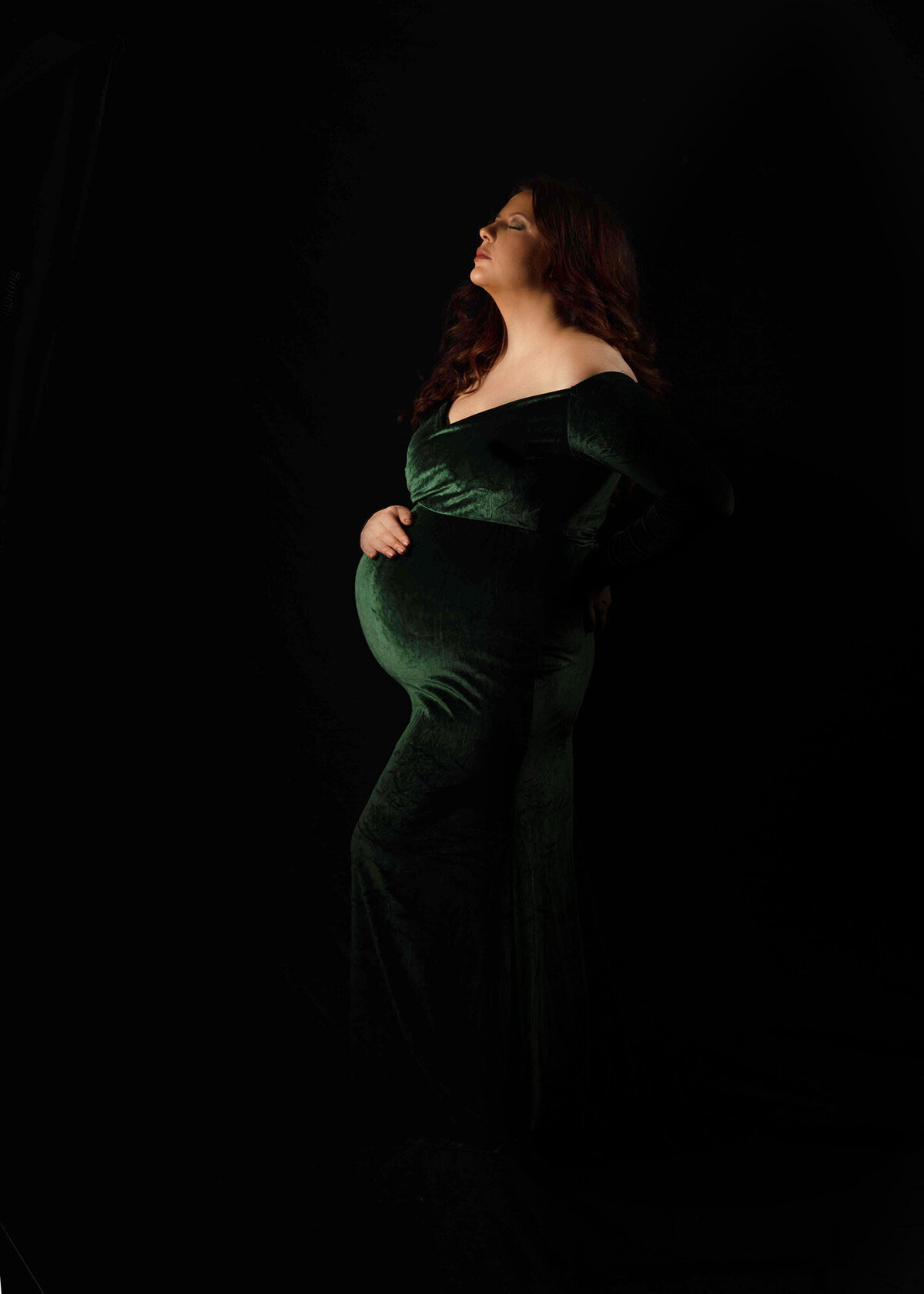 A woman stands with a hand on her bump in a green velvet maternity gown while looking up