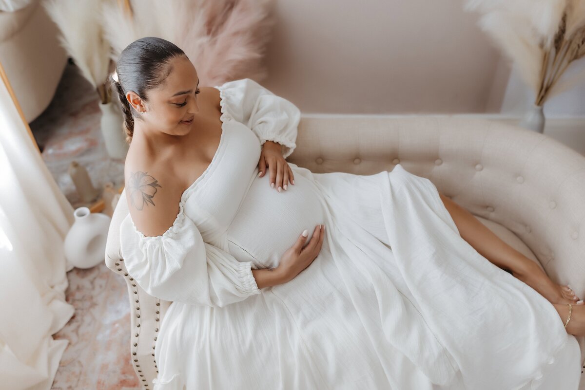 Pregnant woman in a white dress sits on a cream-colored couch, gently holding her belly with one hand, in a softly lit room.
