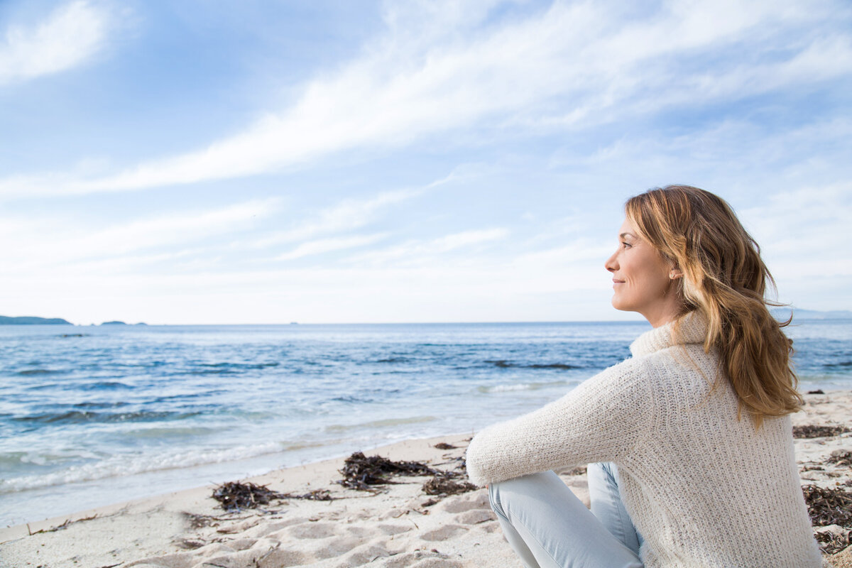 A middle-aged woman sits on the beach and looks off into the distance.