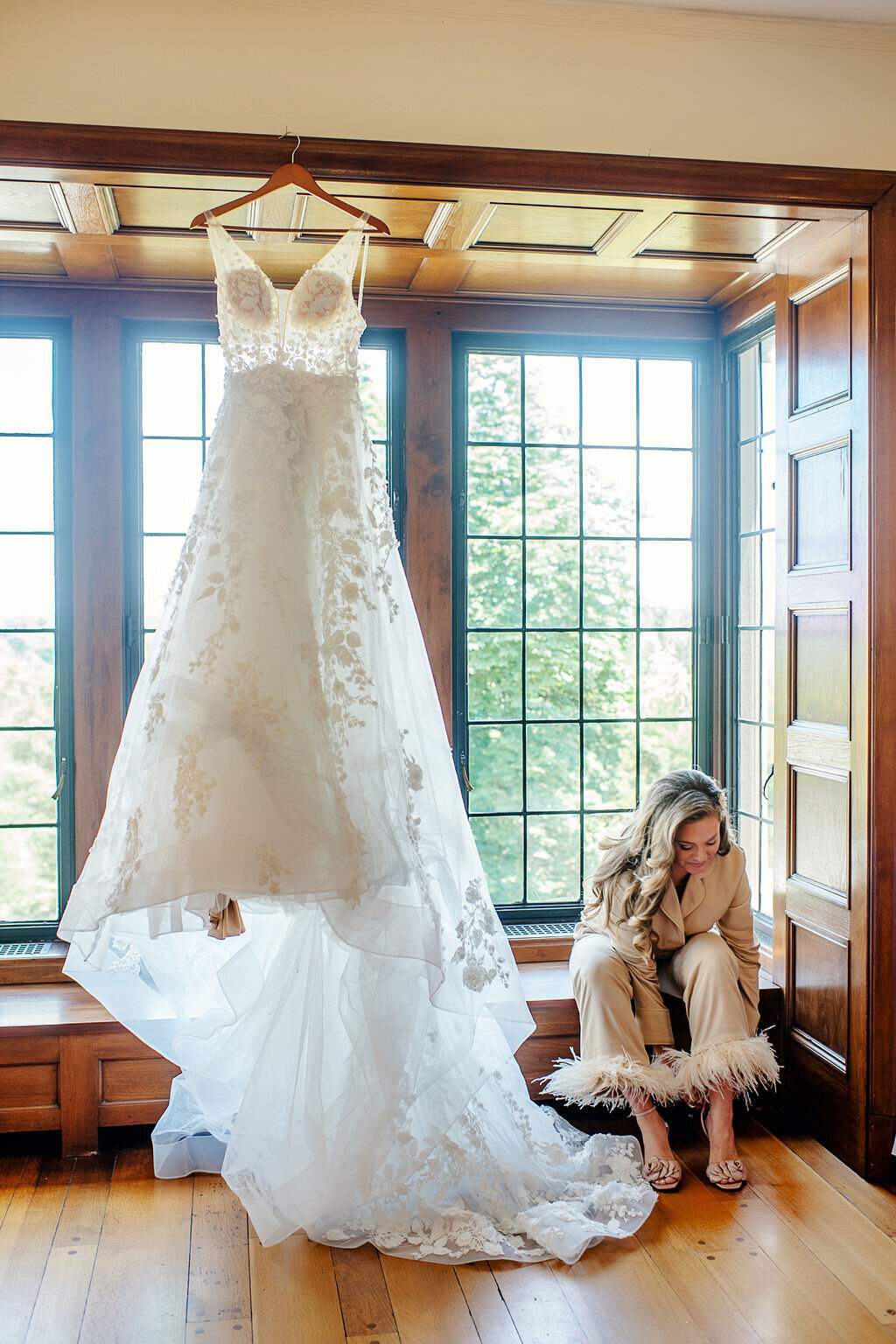 Wedding dress hanging in front of large windows while a woman sits nearby adjusting her shoes.