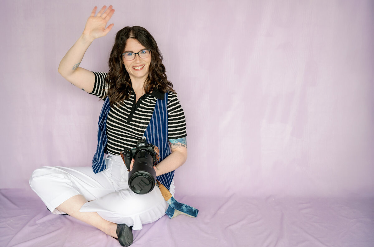 Alicia, lead photographer and owner of Alicia LaPierre Photography, smiling and waving at the camera against a light purple backdrop