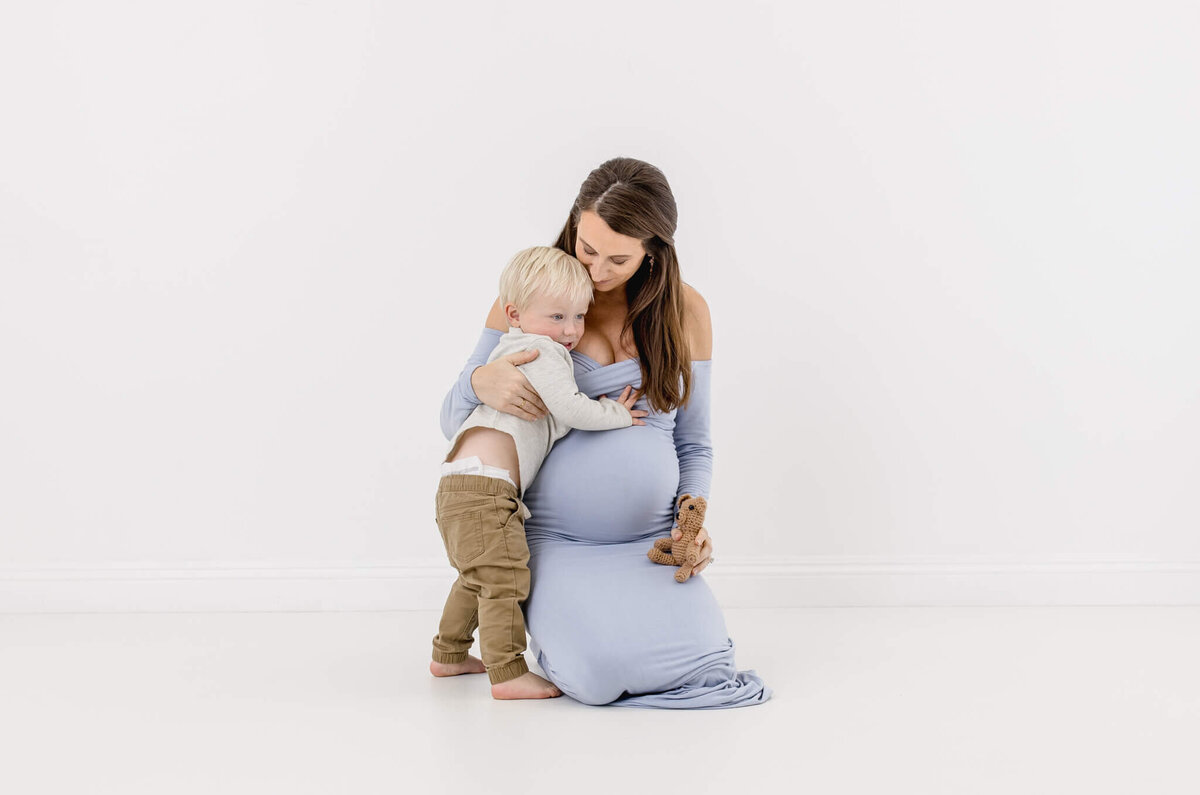 A pregnant woman in a light blue dress kneels on the floor, gently hugging a young boy with blonde hair wearing a light gray sweater and brown pants. The boy holds a small toy in his hand. They are in a bright, white room.