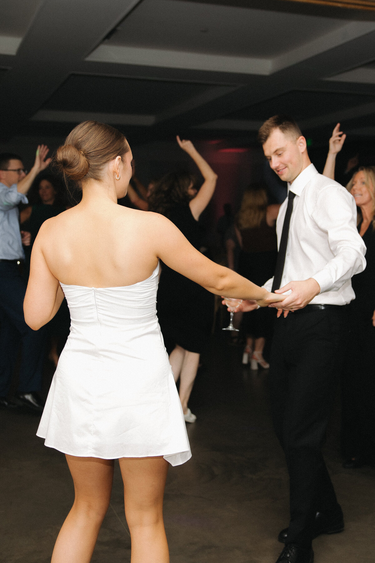 Bride in short, strapless reception dress holds her groom's hand as they face each other dancing. The groom smiles holding a wine glass in the hand that isn't being held. He is in a white dress shirt, black slacks and a thin long black tie. People dance with their hands in the air behind them. 