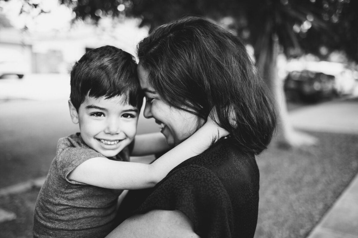 Black & white image by Los Angeles photographer Marjorie Cohen of a mom holding her toddler son as he smiles towards camera.