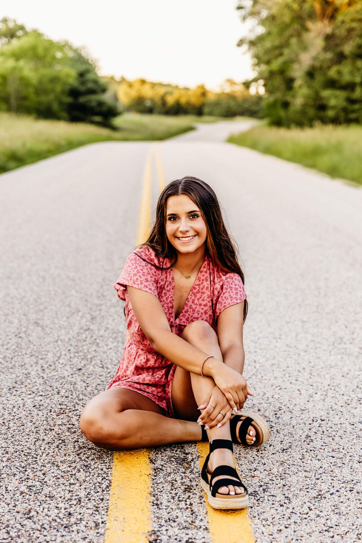 beautiful brunette teenage girl in a pink floral romper sitting with one knee up and the other tucked under as she rests her hands on her knee and looks up while sitting on a backroad near Oshkosh