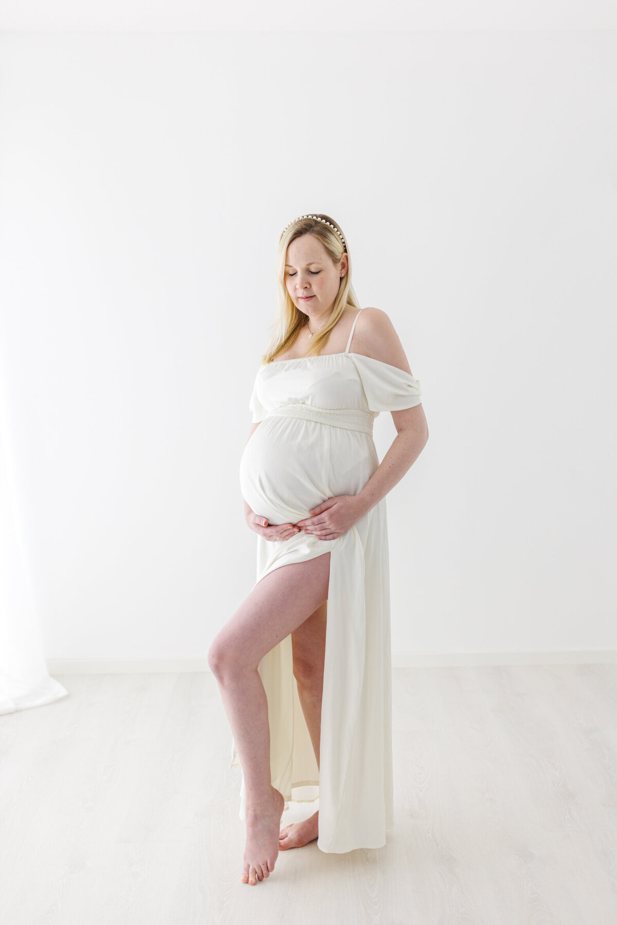 Helprofil av en gravid kvinne i hvit kjole. Hun står med venstre ben løftet, kjolen har splitt og man kan se begge beina. Hun har øynene lukket. Fotografert i Studio Landmark, Fyllingsdalen.