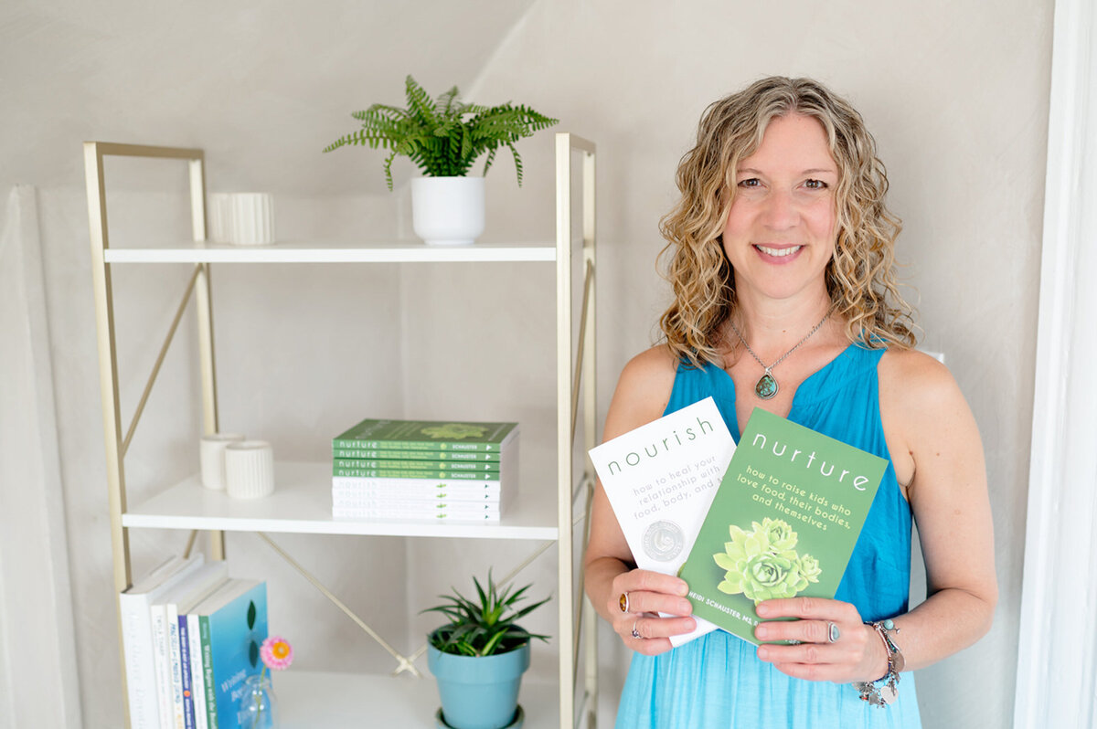 Heidi Schauster in a blue dress holds two books titled "Nourish" and "Nurture" beside a shelf with plants and more books, conveying a sense of calm and knowledge.