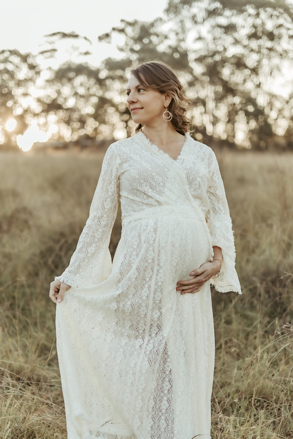Pregnant woman in a lovely white lace robe smiling and holding her baby bump in a beautiful grassy outdoor location