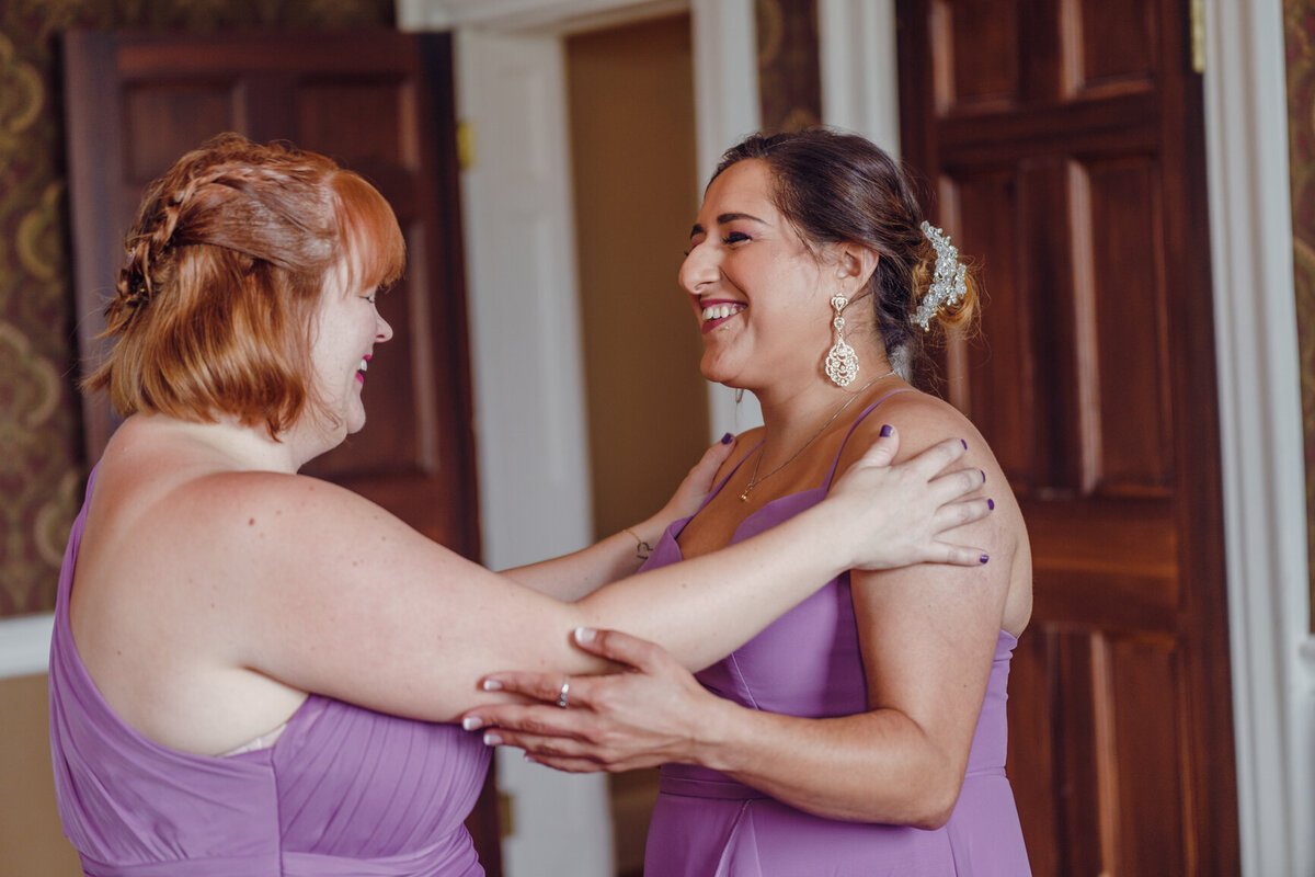 Two women dressed in purple bridesmaids gowns share a laugh  before ceremony at Belmont Country Club in Virginia