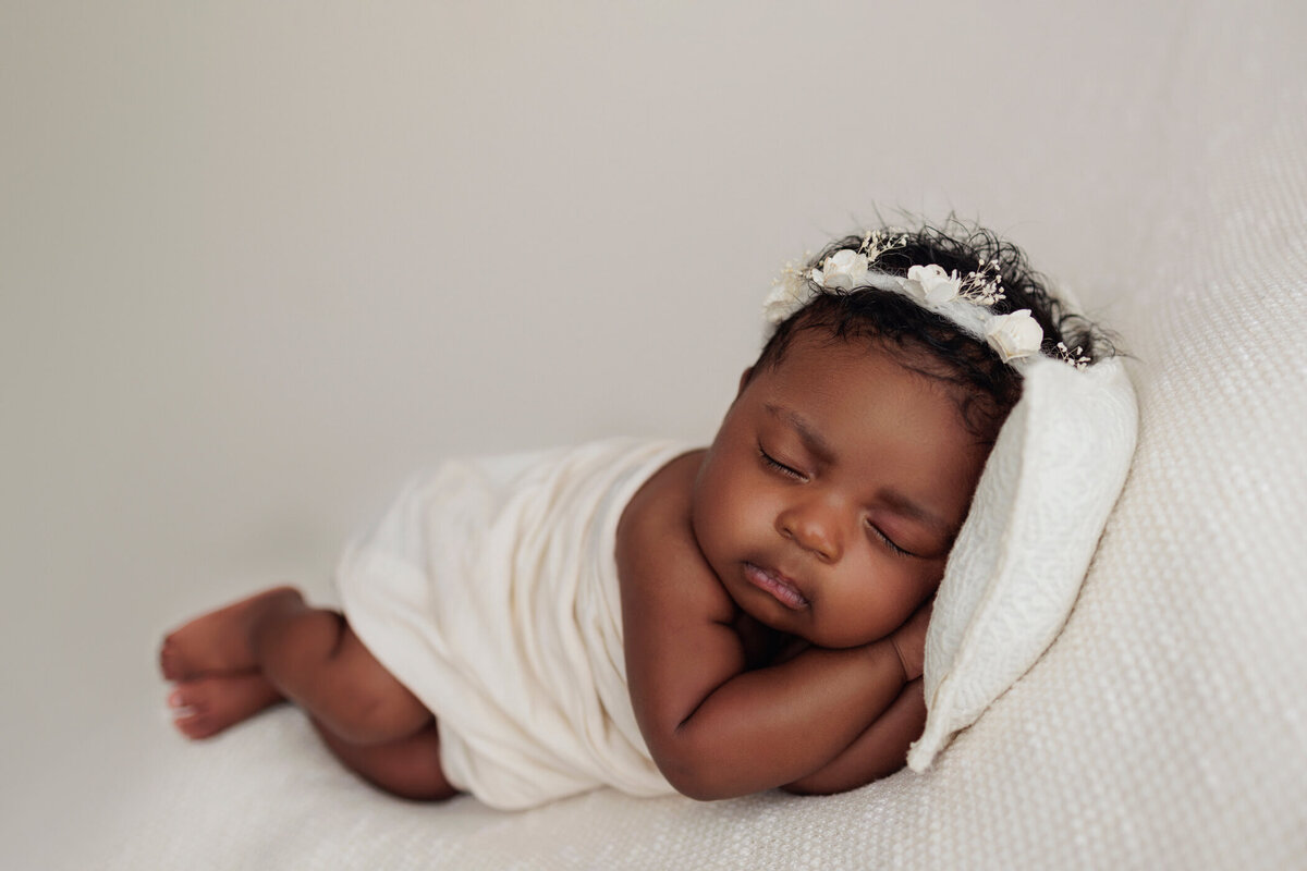 Sleeping newborn baby girl wrapped in a soft white blanket with a delicate white headband, resting on a white pillow on a white backdrop.