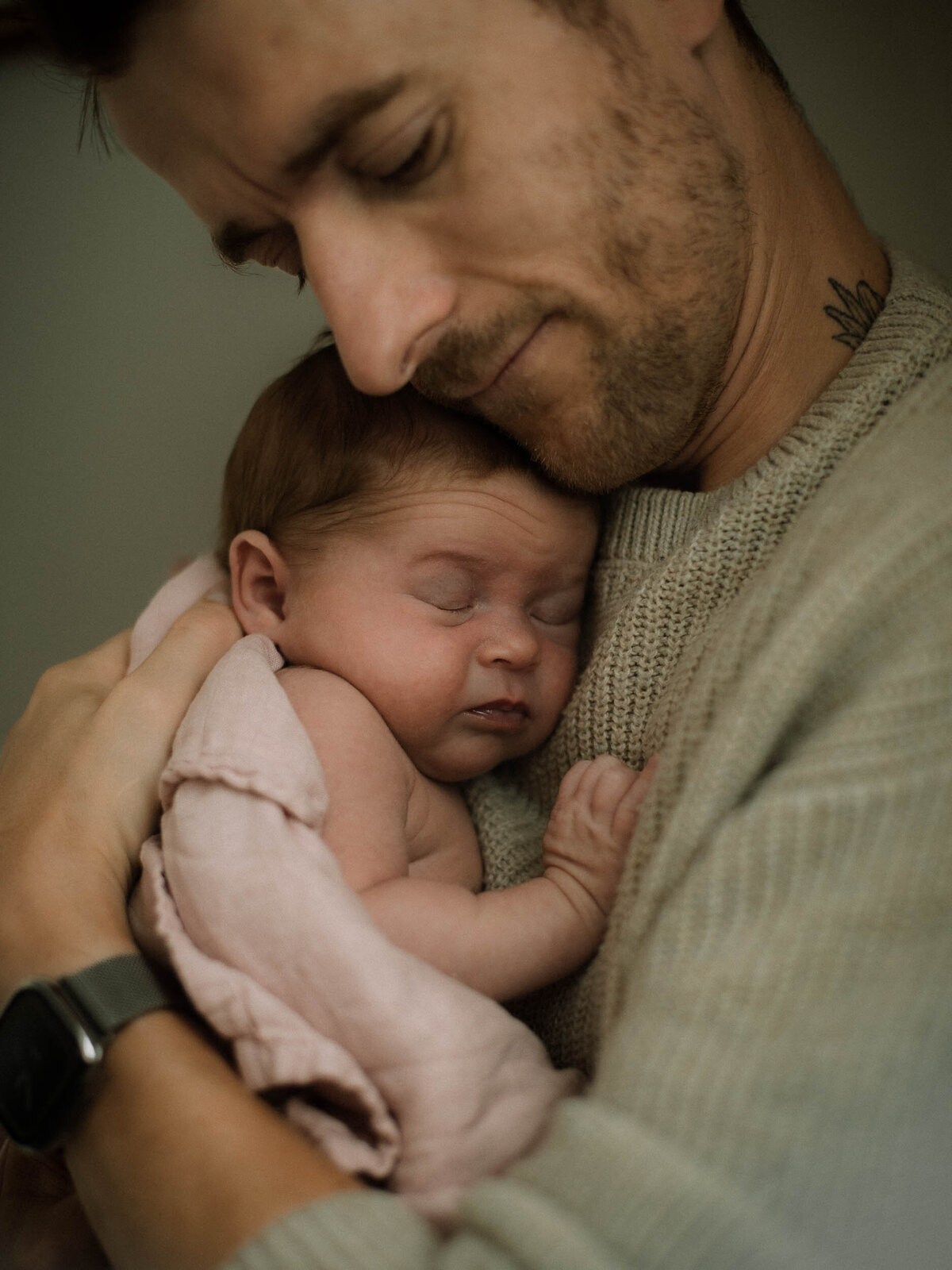 Dad cradling baby girl against his chest while she sleeps and is wrapped in a light pink blanket.