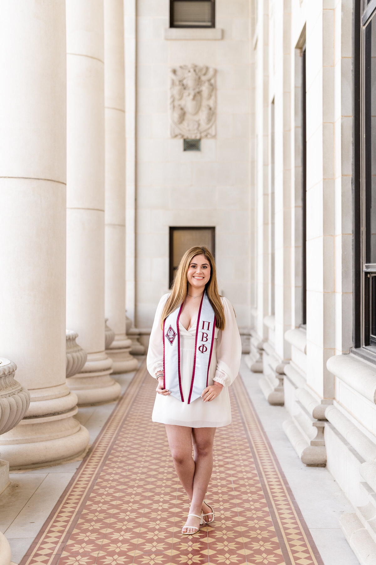 Texas A&M senior girl holding Pi Phi stole while wearing white dress and smiling in the columns of the Administration Building