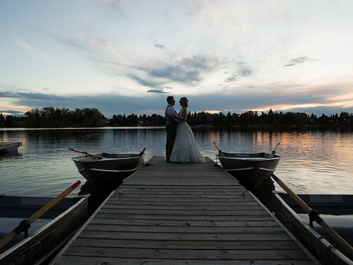 Bride and groom standing on the dock at sunset at The Lakehouse, a romantic sophisticated wedding venue in Calgary, featured on the Brontë Bride Vendor Guide.