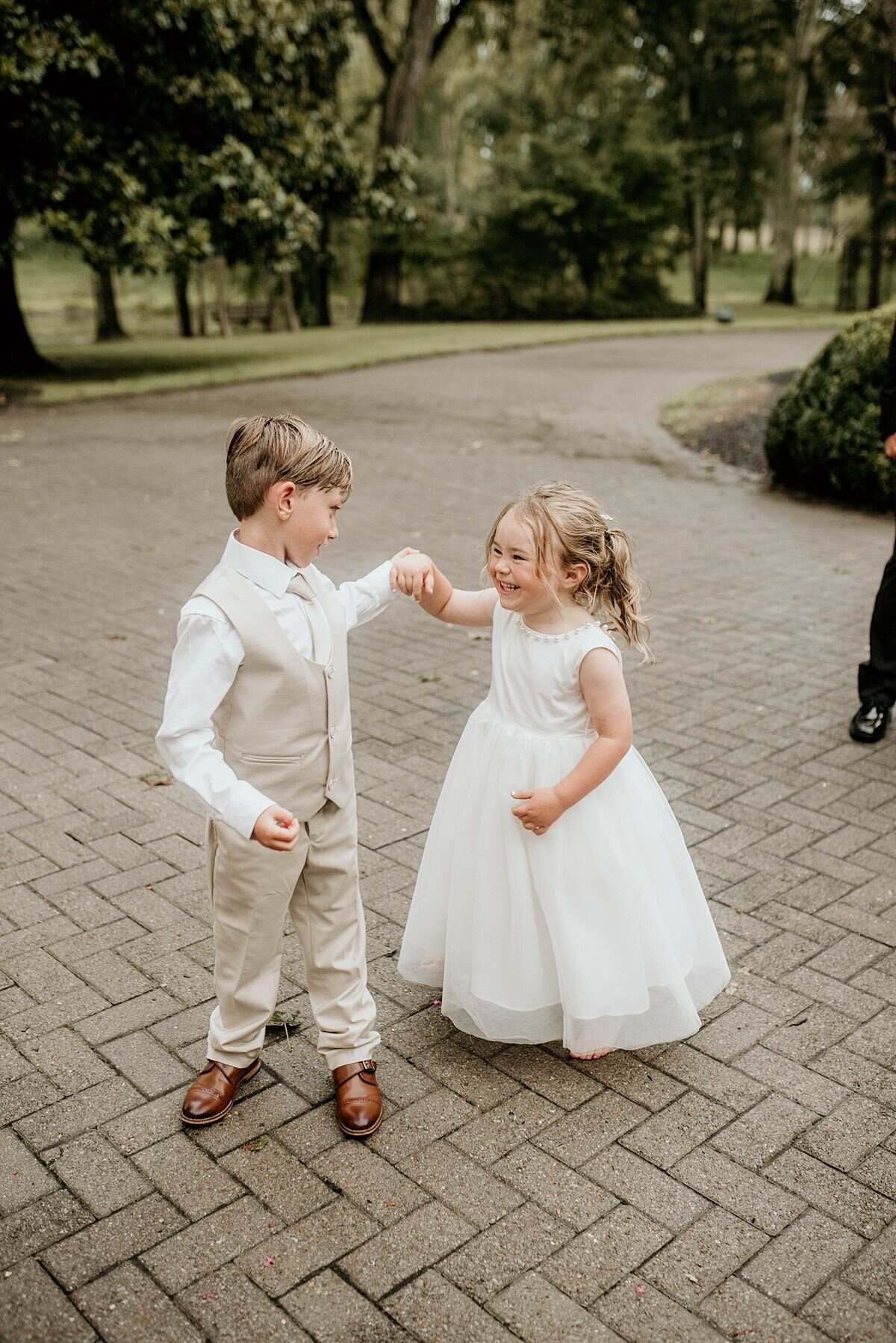 A ring bearer in a white shirt and tan suit dances with the flower girl dressed in a floor length white dress.