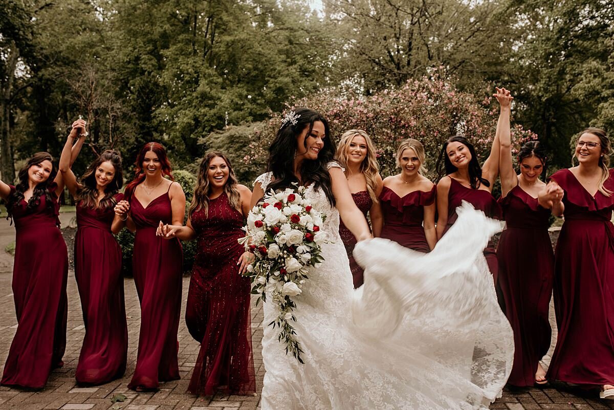 Bride holding a large cascade of red and white flowers as she flips her long tulle wedding gown. Bridesmaids wearing burgundy dresses stand behind the bride and cheer.