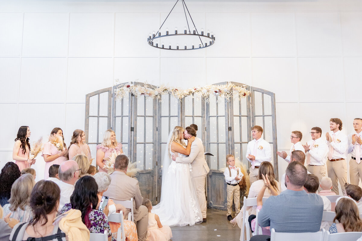 The newlyweds share their first kiss in front of family and friends, standing beneath a beautifully decorated altar. Bridesmaids, groomsmen, and guests clap and cheer as the couple begins their life together.