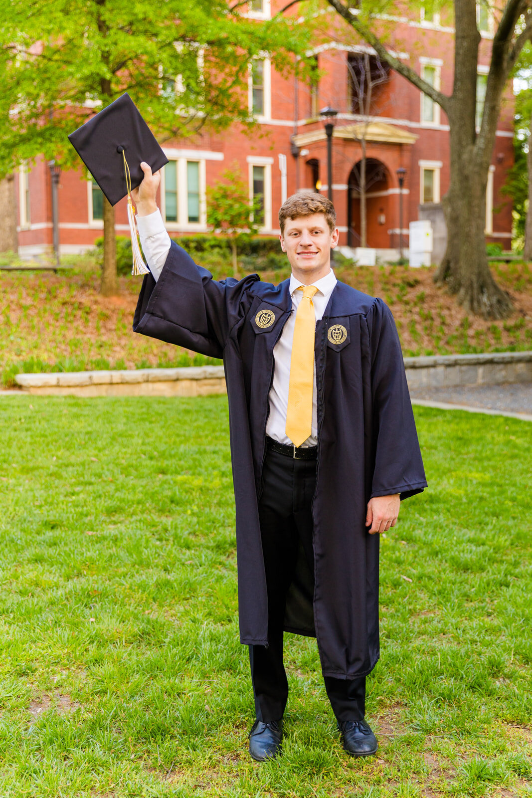 Senior boy wearing a costume white shirt and yellow tie and graduation outfits cap and gown standing one hand holding the cap in the air ion campus during graduation photos with Atlanta photographer Laure Photography