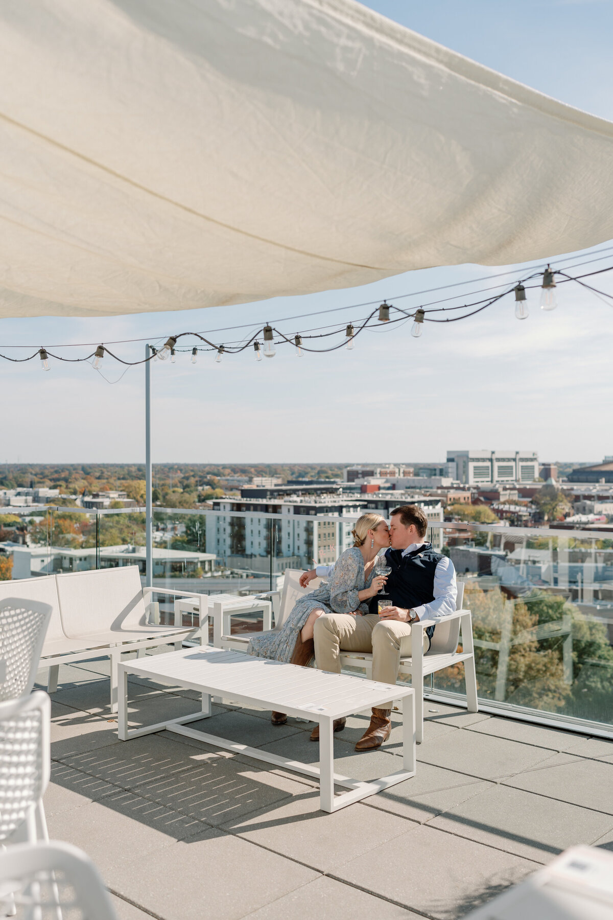 The Quirk Hotel Rooftop Engagement Photos | couple sharing a kiss while sitting on the white rooftop patio furniture of the Quirk Hotel with views of downtown Richmond around them