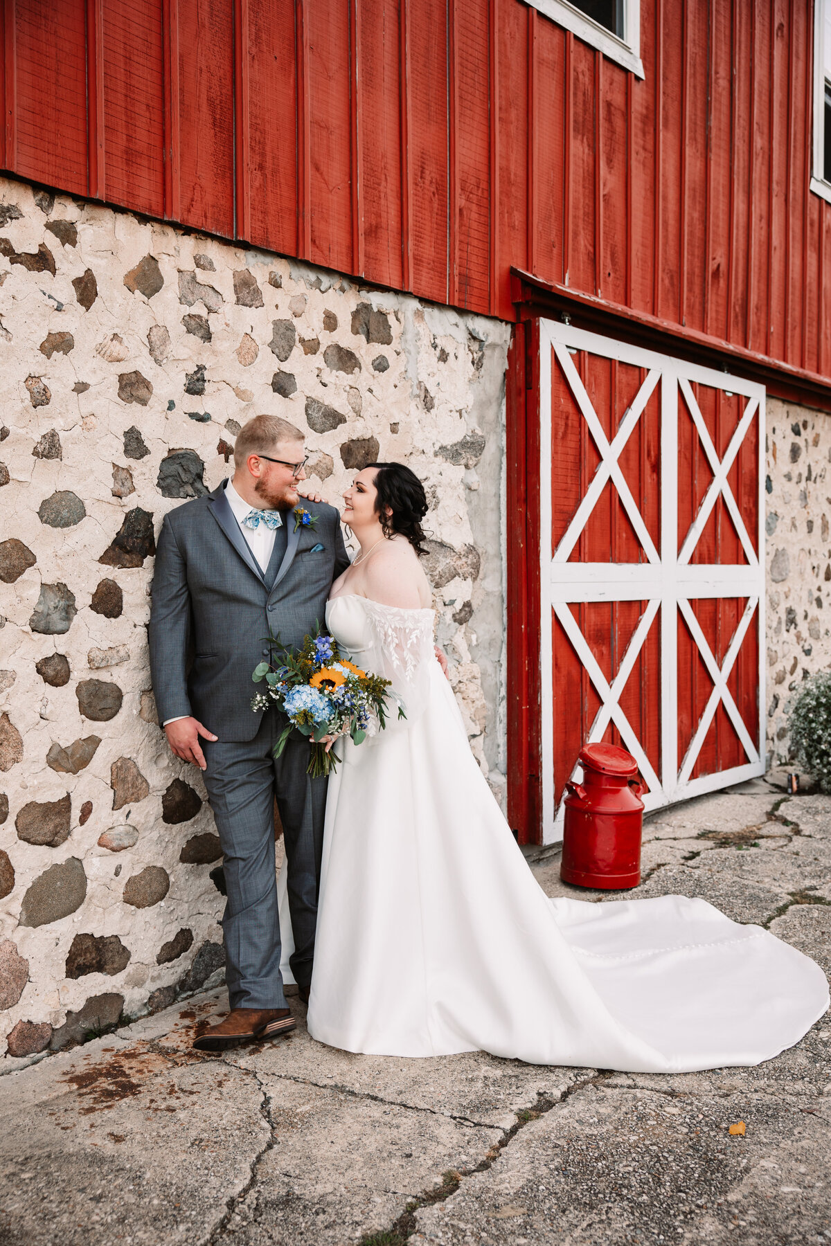 A groom leans on a stone barn wall with his bride leaning on him