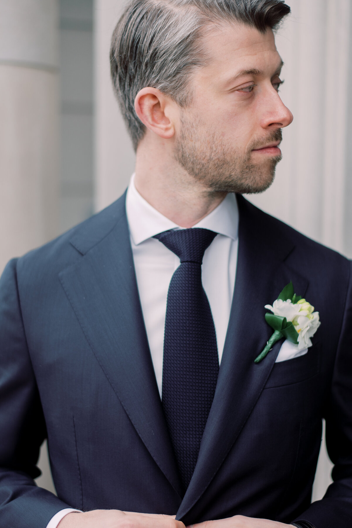 A sophisticated close-up portrait of a groom adjusting his suit jacket, showcasing a classic boutonniere of white flowers against a dark navy suit
