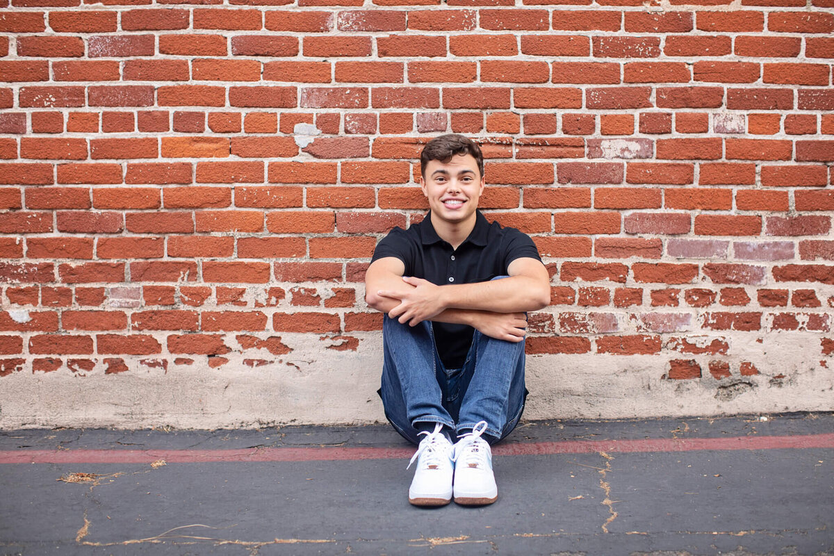 A young man sits on the ground in front of a red brick wall, smiling. He wears a black shirt, jeans, and white sneakers.