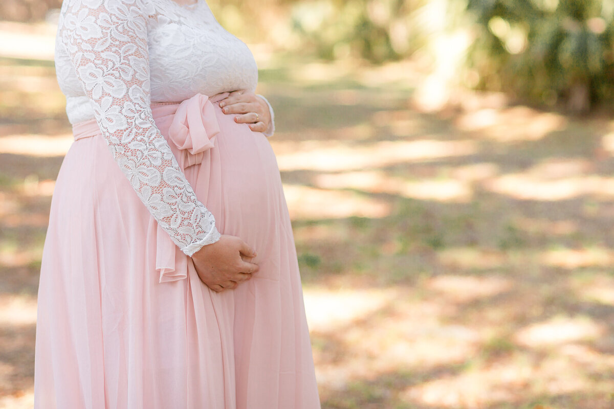 New young mom wearing a lace shirt and pink skirt holding tummy at Wormsloe in Savannah, Ga