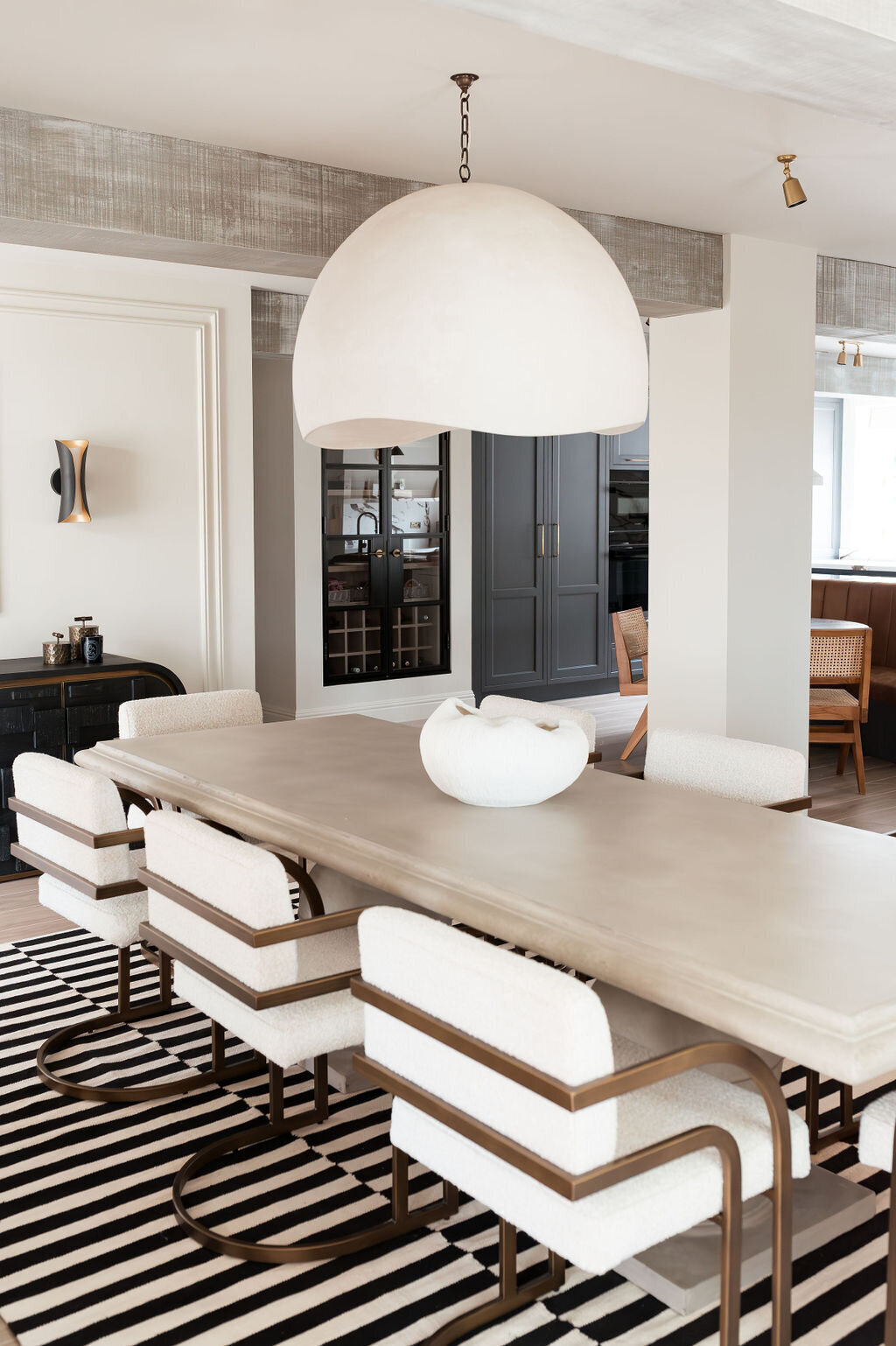 A modern dining room with a large rectangular wooden table surrounded by eight cushioned chairs. A large dome-shaped pendant light hangs above the table, and a black and white striped rug lies underneath.