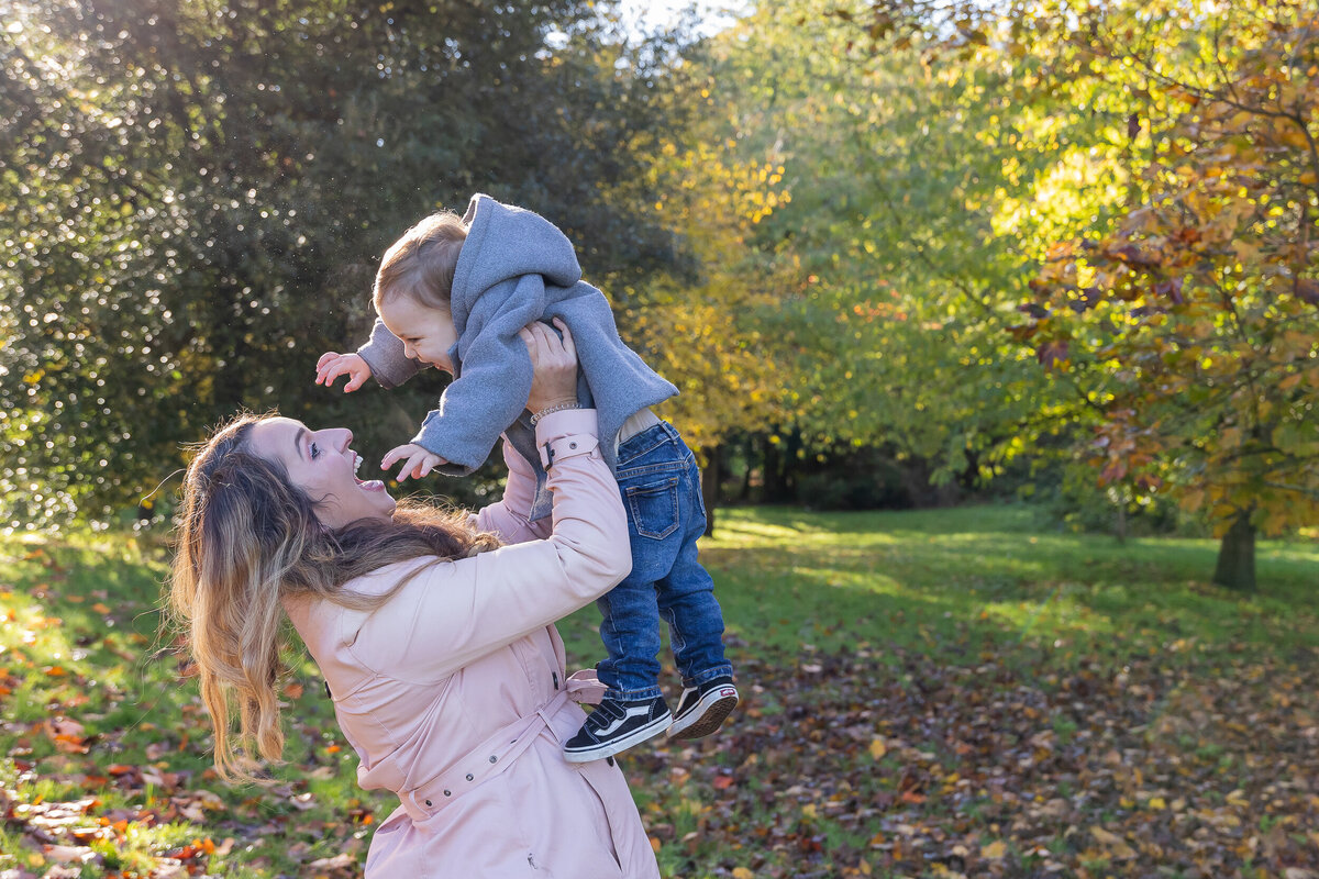 A woman lifts a young child in the air in a sunlit autumn park surrounded by colorful trees and fallen leaves.